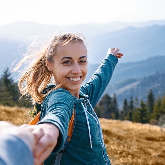 first person view of a happy attractive woman hiker standing on edge of mountain ridge against background of sunset. Woman is holding someone hand and greeting a sunset. Traveling and follow me concept